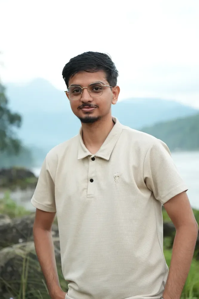 Anuj Paudel in a beige polo shirt and glasses, standing before misty hills and a wide river at British Camp