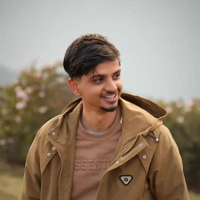 Anuj Paudel smiling in a tan hooded jacket beside the Panch Pokhari trail, with pink blossoms blurred behind