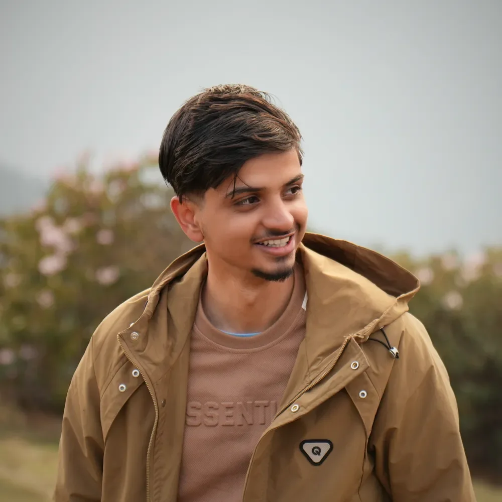 Anuj Paudel smiling in a tan hooded jacket beside the Panch Pokhari trail, with pink blossoms blurred behind