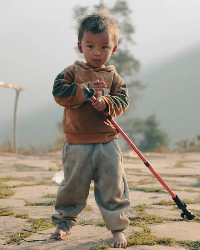 A small barefoot child holds a red trekking pole on a stone path at Panch Pokhari, with hazy hills behind