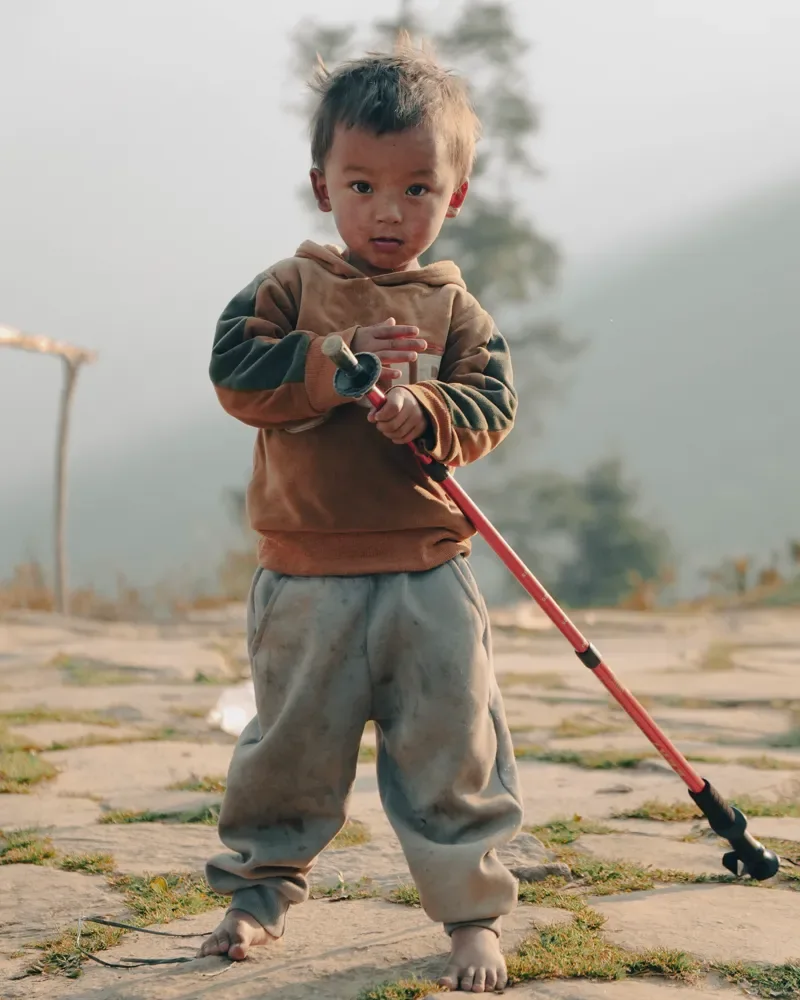 A small barefoot child holds a red trekking pole on a stone path at Panch Pokhari, with hazy hills behind