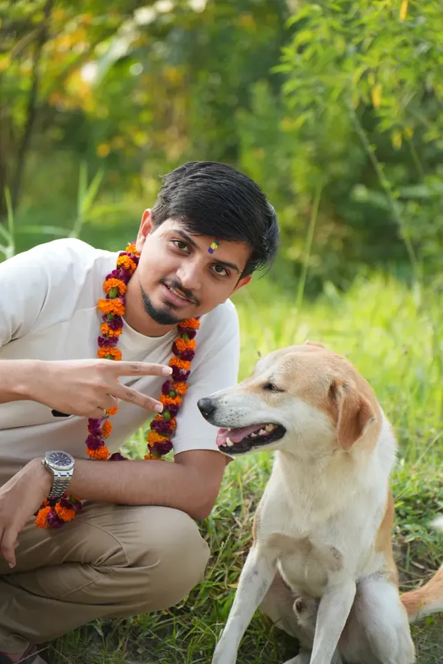 A man in a marigold garland and a small tika crouches beside a seated tan and white dog during Tihar, Nepal