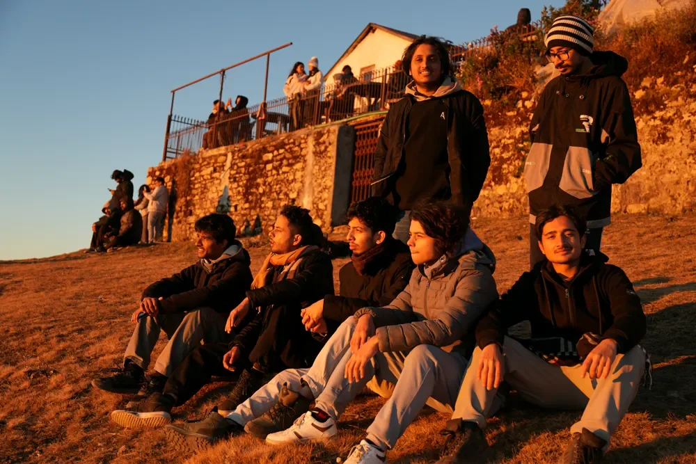 A group of trekkers sit and stand on a dry slope at Sailung in warm low light, with a stone wall behind them