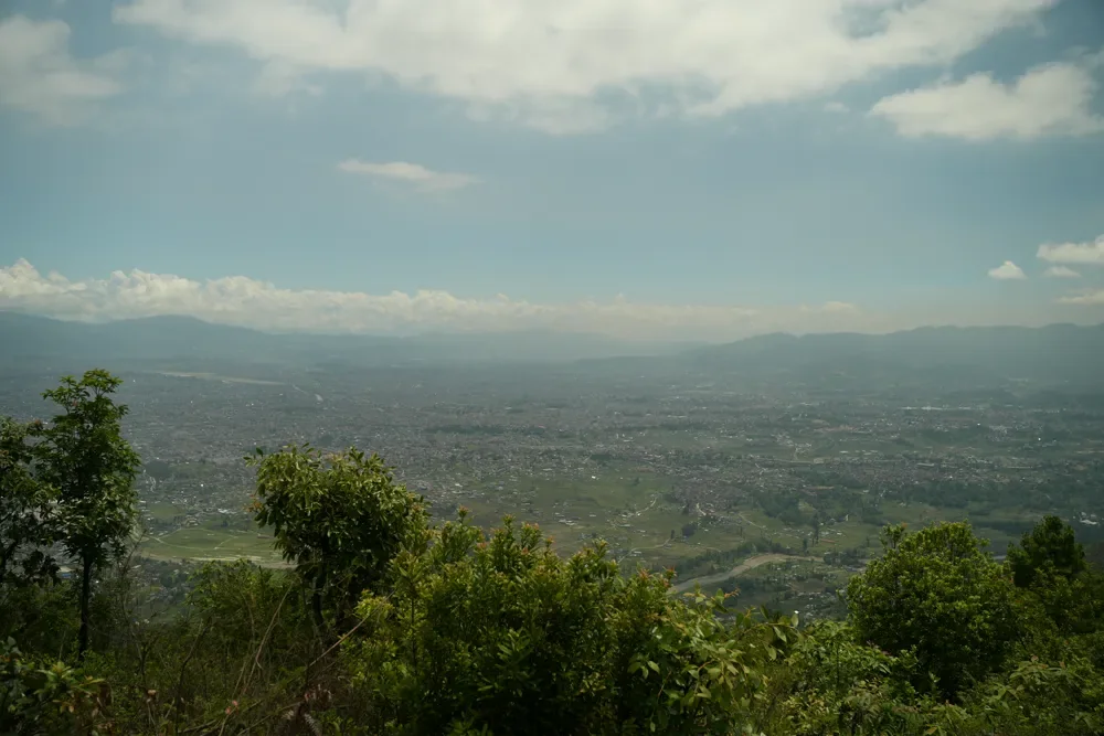 Wide view over a hazy valley of fields and houses from a Champadevi ridge, with hills and clouds beyond
