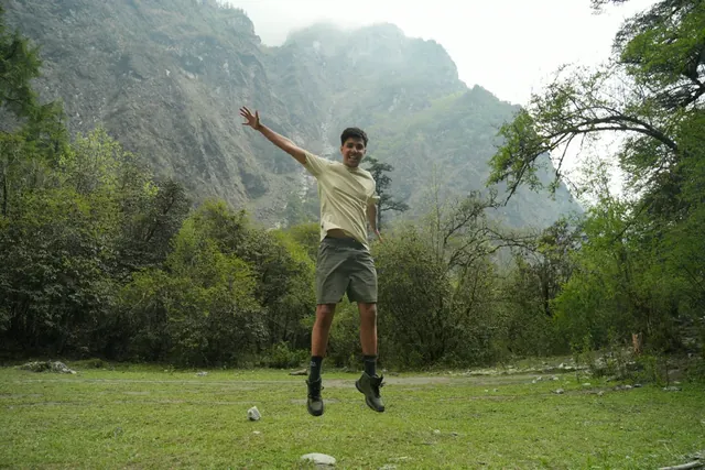 A hiker in a pale yellow T-shirt leaps into the air on a grassy clearing below a steep, misty valley wall in Langtang