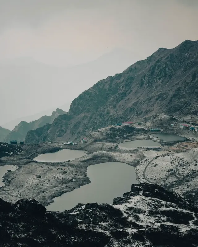 Several small grey lakes in a rocky, snow-dusted basin with huts on the ridge, under a misty sky at Panch Pokhari