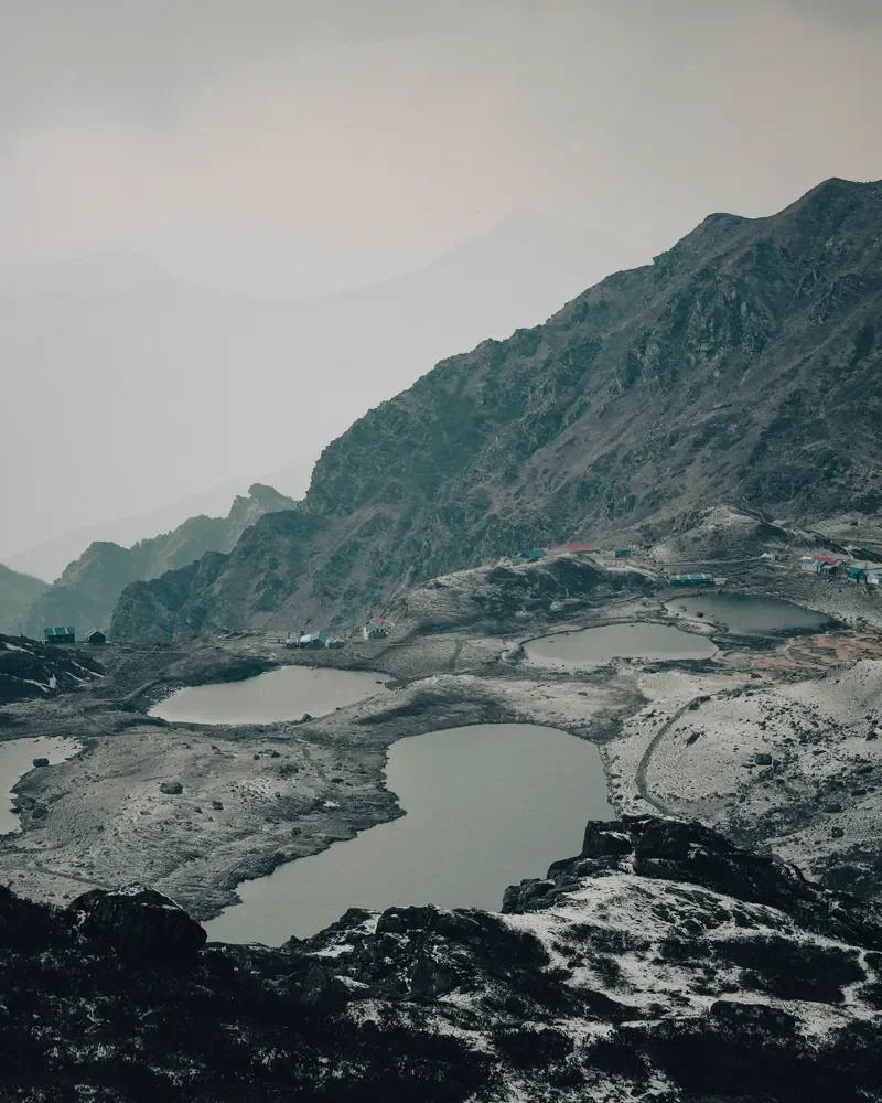 Several small grey lakes in a rocky, snow-dusted basin with huts on the ridge, under a misty sky at Panch Pokhari