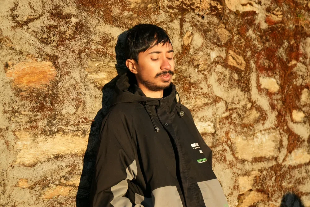 A man with his eyes closed stands against a weathered stone wall in warm, low sun at Sailung