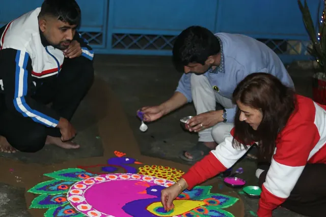 Three people crouch over a peacock rangoli of pink, purple and green powder at night during Tihar, Nepal