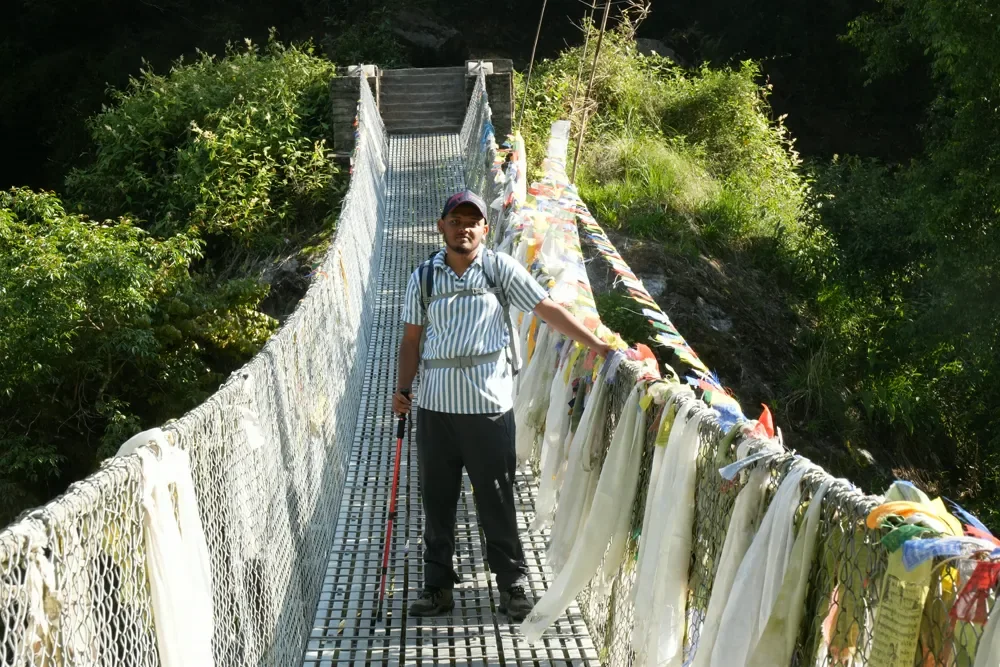 A trekker with a red pole stands on a suspension bridge draped with prayer flags in the Langtang valley