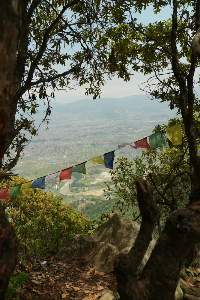 Prayer flags strung between trees on the Champadevi ridge, above a hazy valley of houses and fields