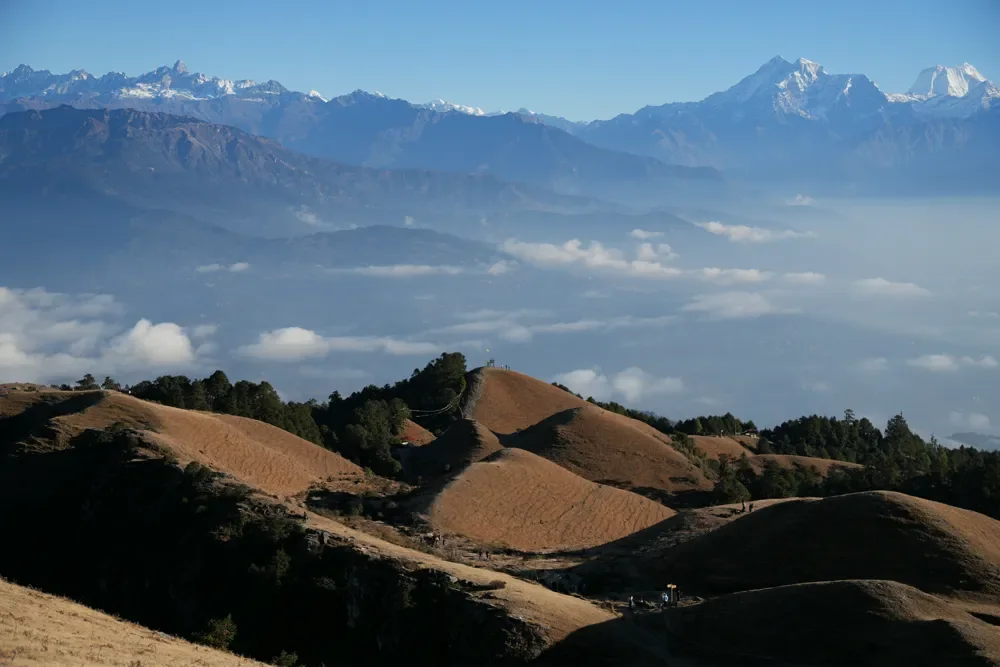 Golden grass hills with a layer of cloud in the valley and a snow-capped Himalayan range behind, at Sailung