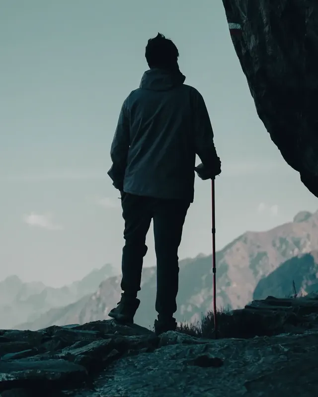 A trekker in silhouette on a rocky ledge with a red trekking pole, facing hazy mountain ridges at Panch Pokhari