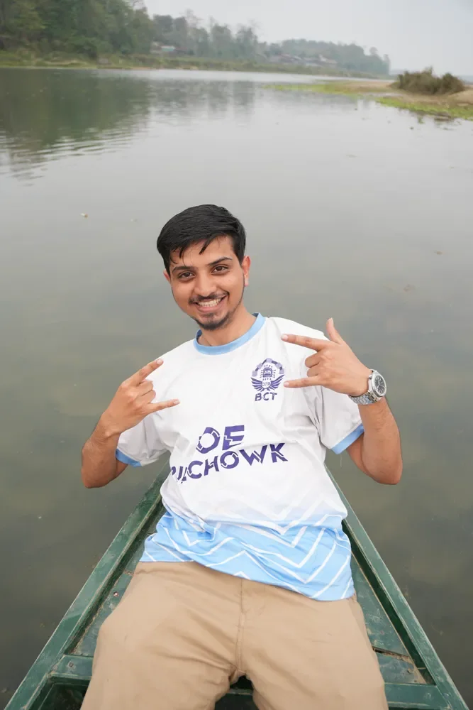 A smiling man in a white and light blue jersey sits in a green wooden boat on calm water, making hand signs, Kathmandu