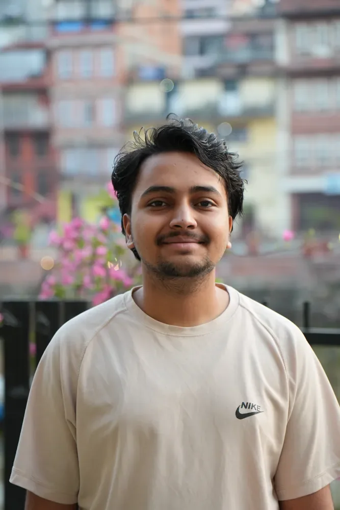 A smiling man in a cream T-shirt stands in front of a black railing, with pink flowers and buildings blurred behind, Nepal