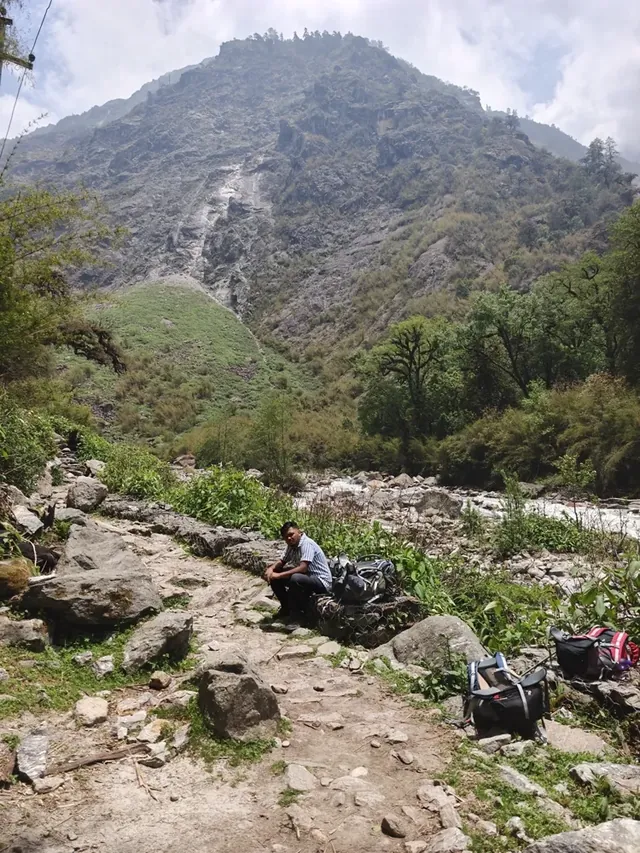 A man sits on a rock by a rocky trail, small against a huge forested ridge above a river in the Langtang valley
