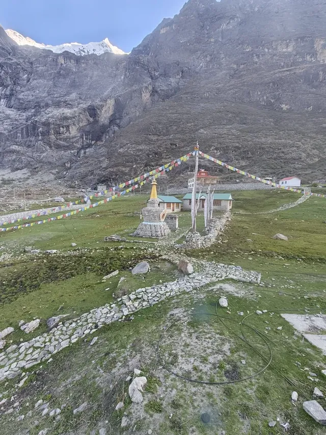 A stone stupa with a golden spire and strings of prayer flags in a green meadow below rocky cliffs at Kyanjin Gompa