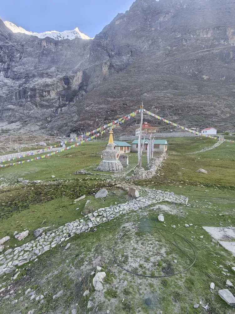 A stone stupa with a golden spire and strings of prayer flags in a green meadow below rocky cliffs at Kyanjin Gompa