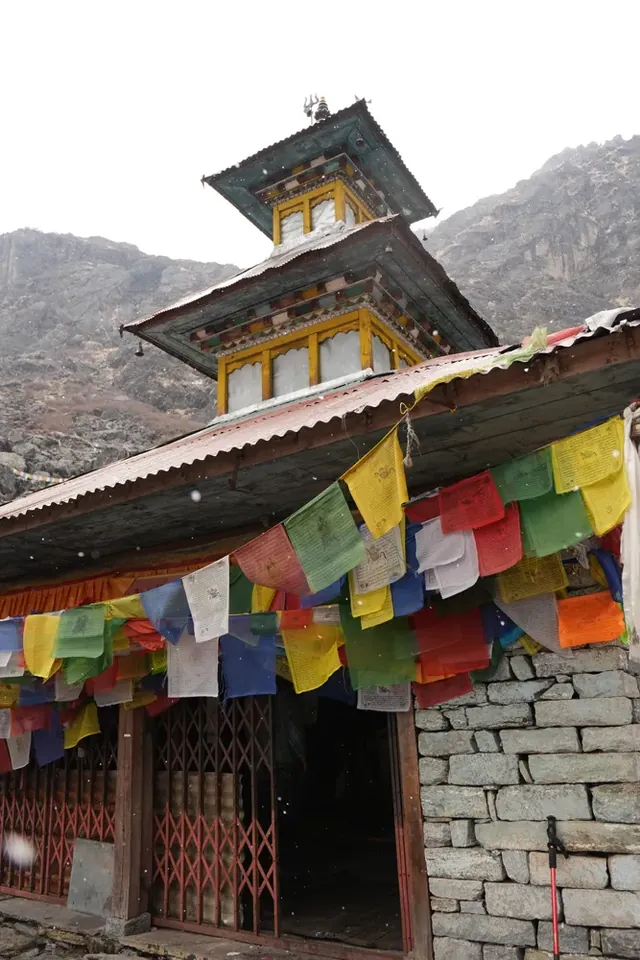 Two-tiered temple with a red lattice gate and rows of prayer flags under light snowfall, Panch Pokhari