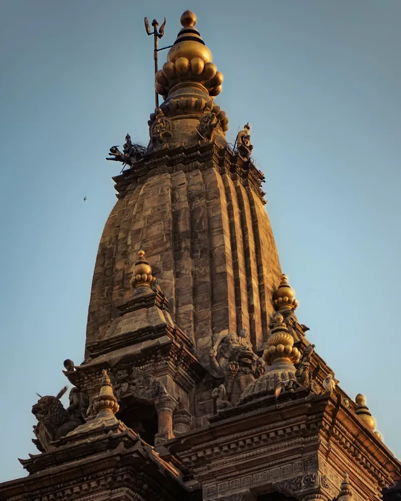 A carved stone temple spire in Kathmandu with gold-capped tiers and a trident on top, in golden light