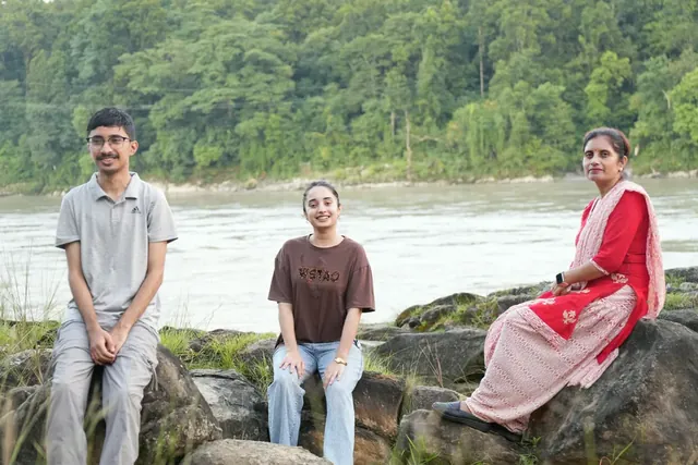 Three people sit on large boulders beside a wide river with a dense green forest on the far bank at British Camp