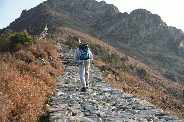 A trekker with a blue backpack walks up a stone-paved path toward a rocky ridge and prayer flags at Panch Pokhari