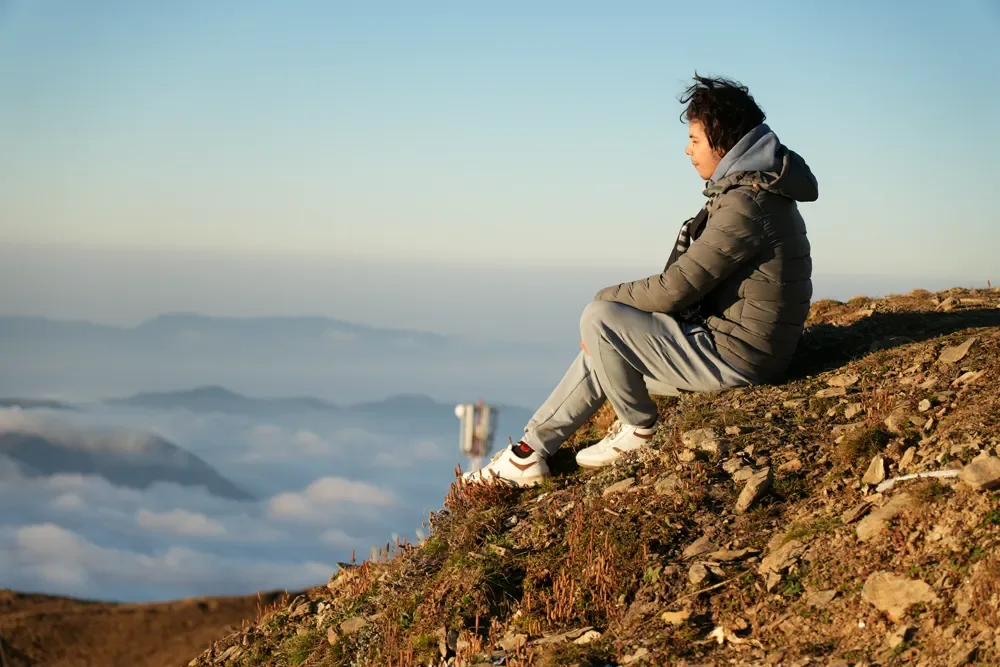 A trekker in a grey puffer jacket sits on a hillside in golden light, looking over a sea of clouds at Sailung
