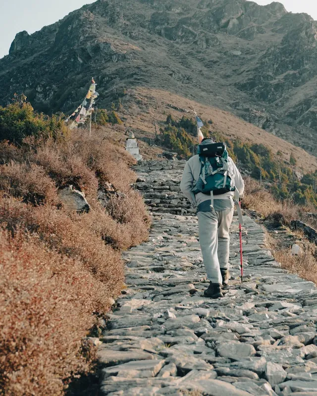 A trekker with a teal backpack climbs a stone path past prayer flags and a small stupa at Panch Pokhari