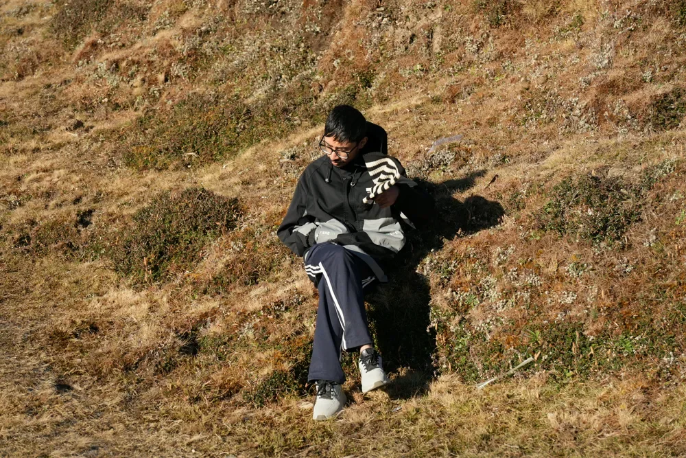 A trekker in a dark jacket and navy track pants sits on a dry, sunlit grassy slope, head bowed, Sailung