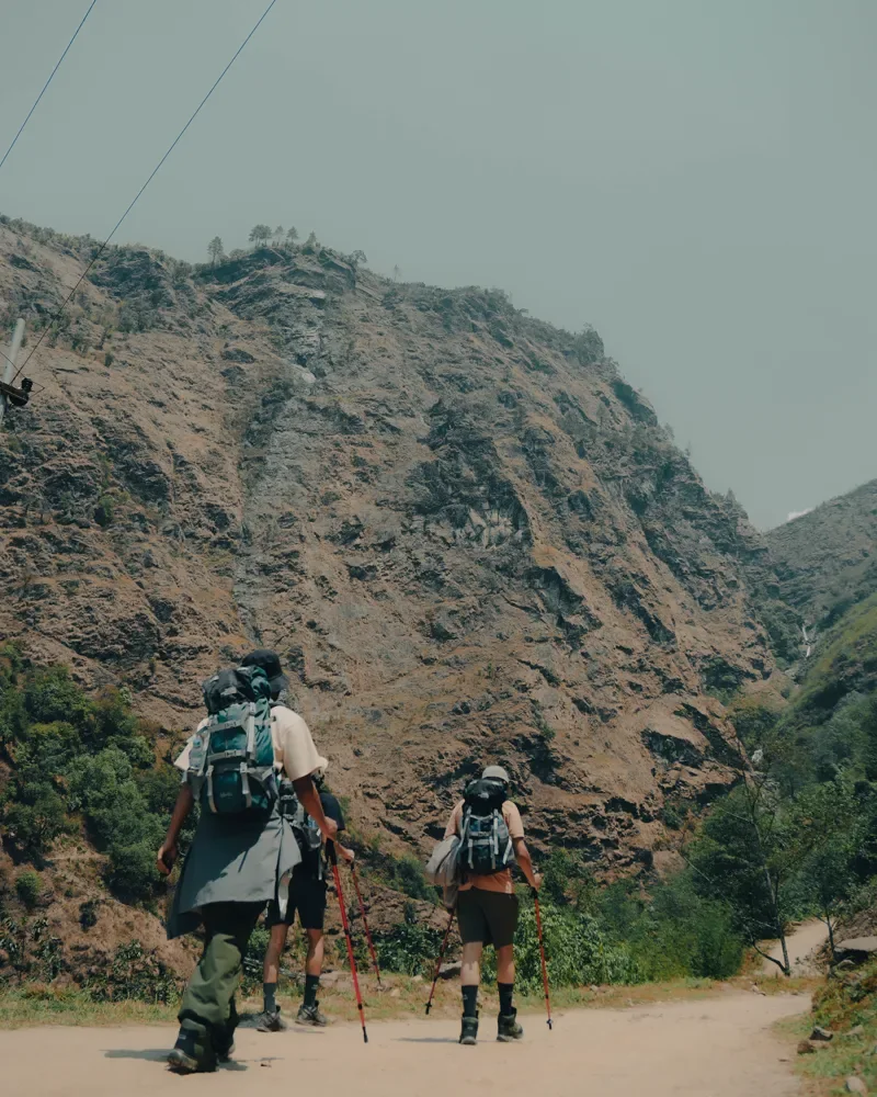 A group of trekkers with backpacks and red poles walk a dirt track below a steep, dry hillside, Panch Pokhari