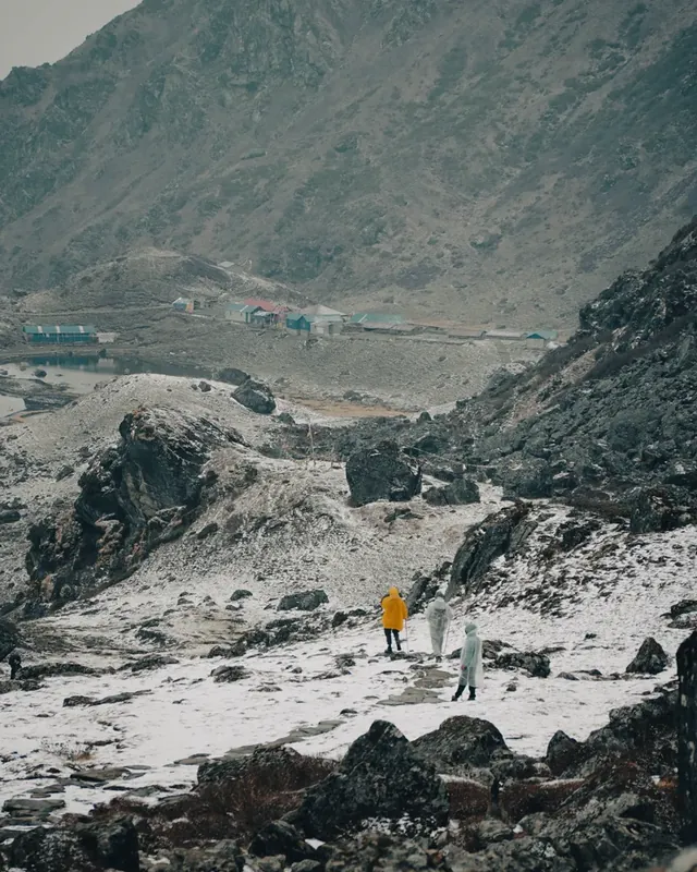 Trekkers in rain ponchos walk along a snowy path toward a small lake village at Panch Pokhari