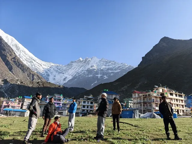A group of trekkers on a grassy flat in front of village buildings and a snow-covered peak in the Langtang valley