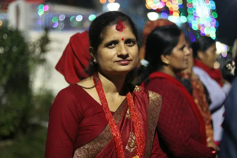 A woman in a maroon sari with a red tika at night, with blurred green, orange and pink Tihar lights behind her